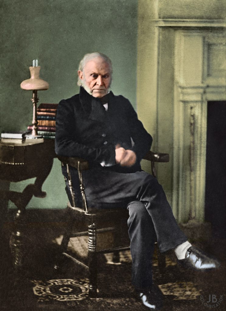President John Quincy Adams having his portrait taken in a study with a table, lamp and books behind him. Adams is sitting on a chair holding his hand and a fireplace is to the right of him