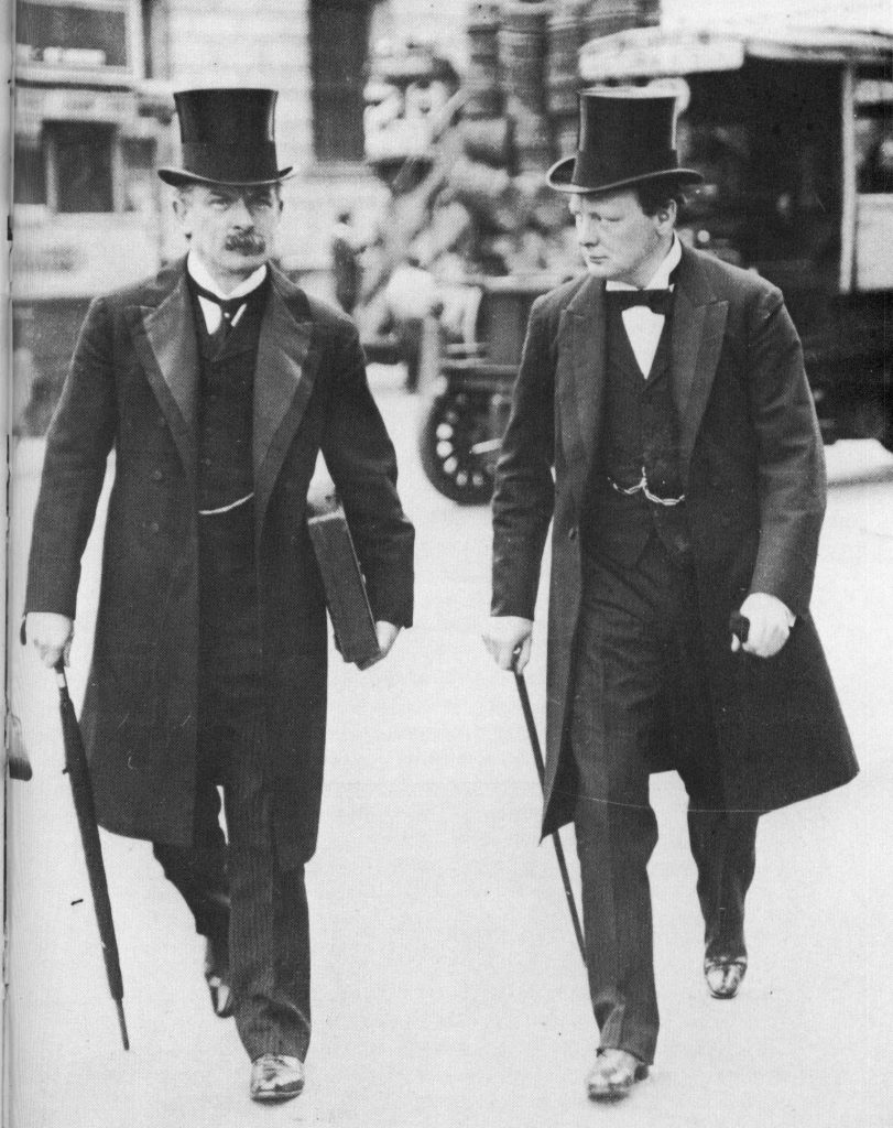 David Lloyd George and Winston Churchill photographed together in a suit and top hat with Churchill holding a cane and Lloyd George holding an umbrella and a box. 1907