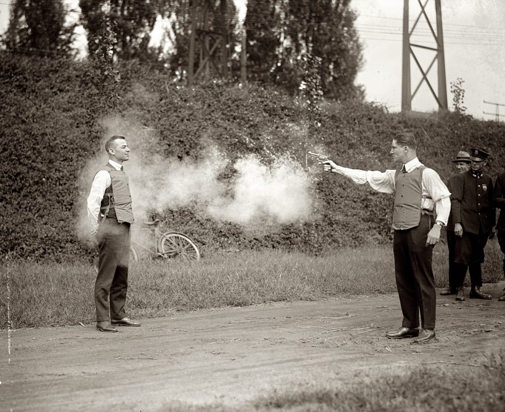 A man aiming a gun at another person wearing a bulletproof vest. Smoke from the barrel surrounds them and onlookers watch on. September 1923.