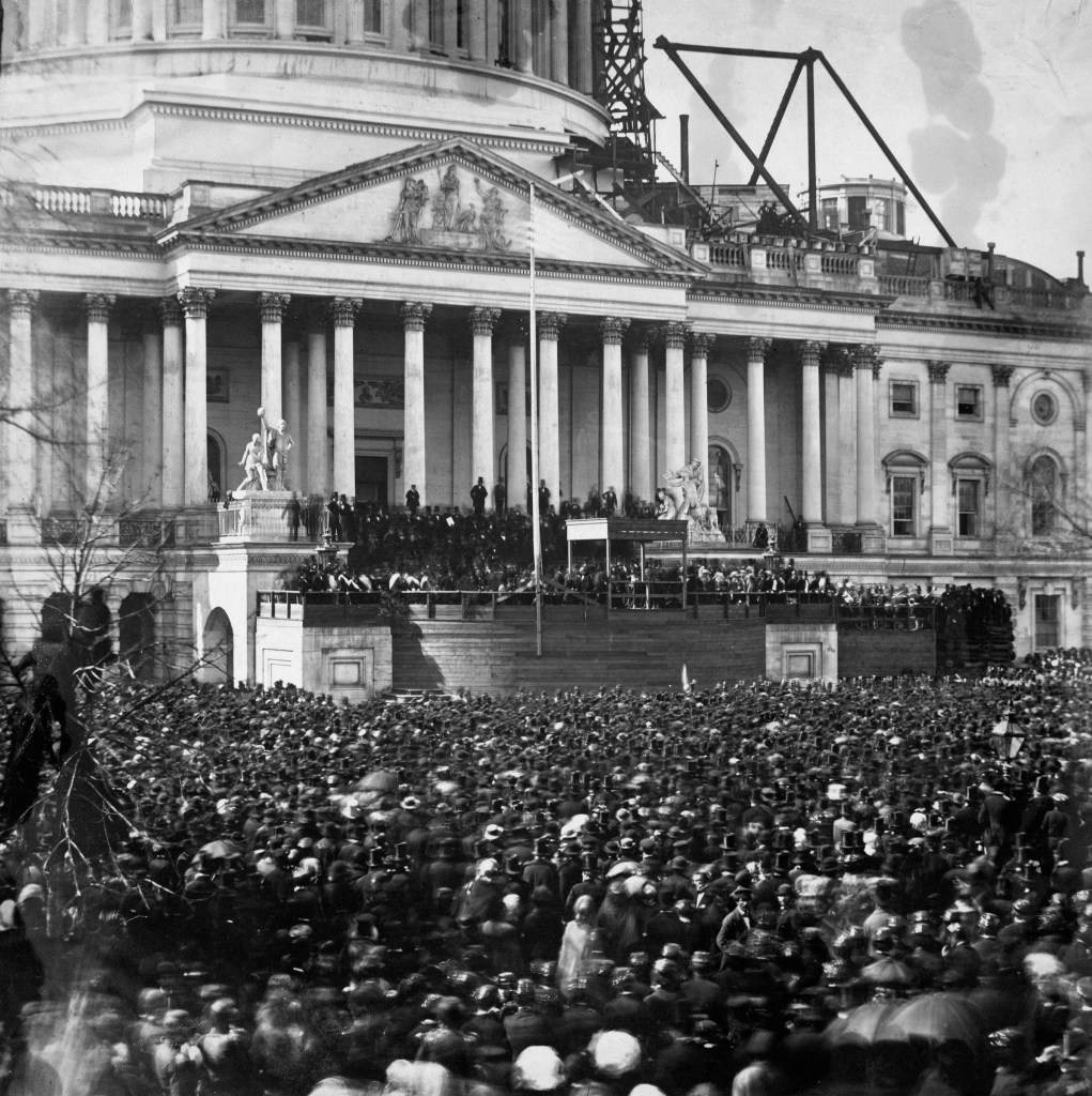 A photograph of the first inauguration of Abraham Lincoln. Thousands of people are crowded around the capitol building in Washington, D.C. that is under construction in 1861.