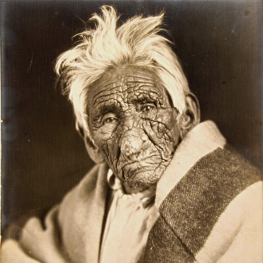 Chief John Smith with white hair and heavily wrinkled skin sitting for a photo with a blanket around him in circa 1915