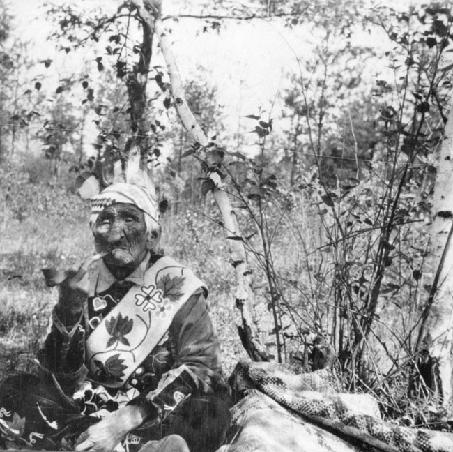 White Wolf Chief John Smith smoking a pipe sitting in a forest while wearing traditional clothing in circa 1910s.
