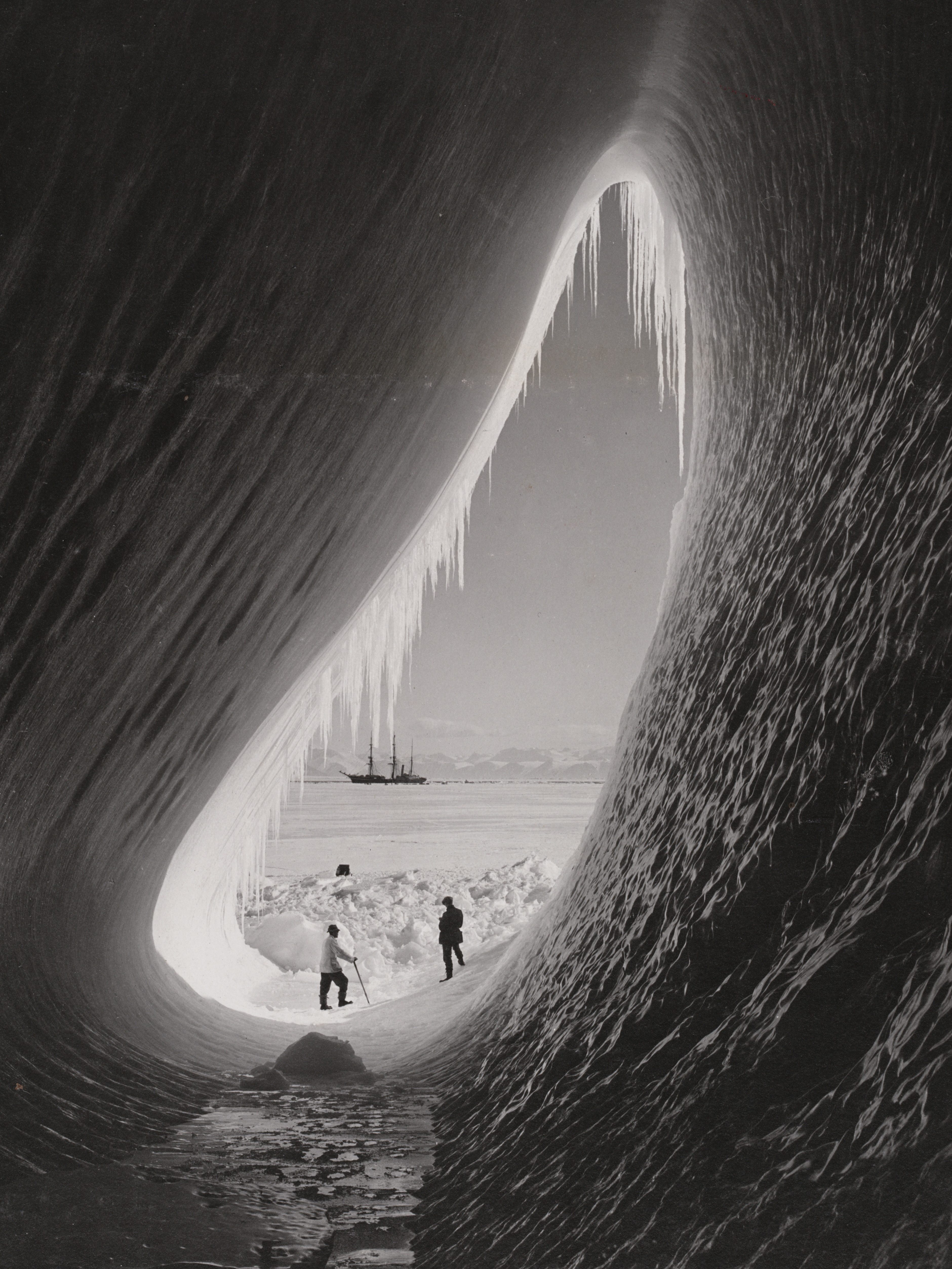 Photograph of two men standing in icy grotto within an iceberg during the Terra Nova expedition with the ship in the background in 1911.