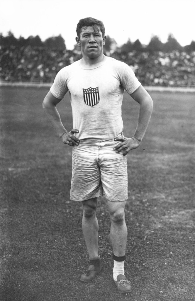 Jim Thorpe pictured in shorts, odd socks and his hands on his waist at the 1912 Olympics.