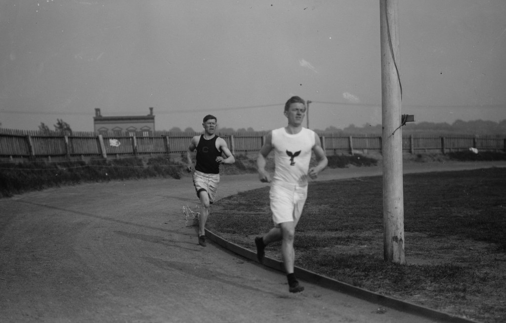 Jim Thorpe running on a track with another man named McGloughlin in Celtic park new york. Thorpe is wearing white shorts and a black vest with a C on it.