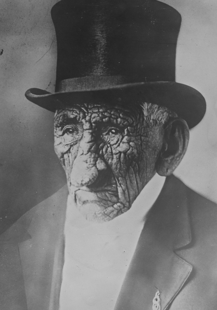 White Wolf Chief John Smith pictured wearing a top hat and suit. You can clearly see his heavily wrinkled face in this image.