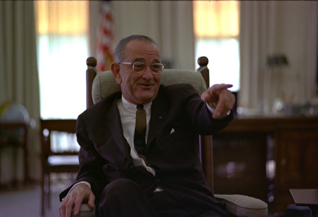 President Lyndon B Johnson sitting in a rocking chair in the White House pointing with his left hand while wearing glasses and having a smile on his face.