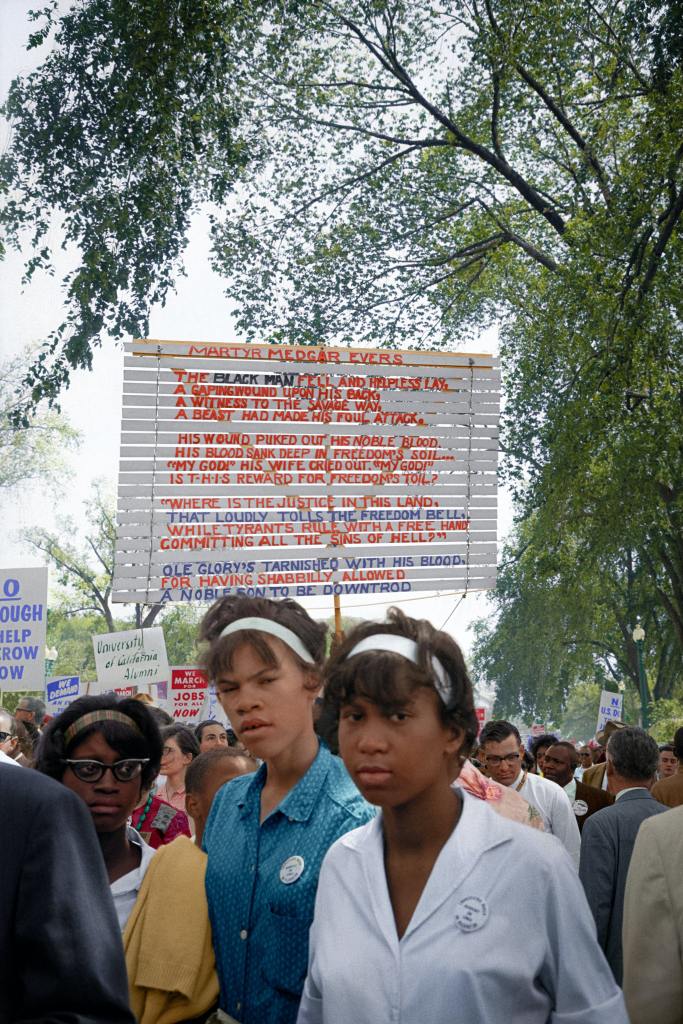 Young women photographed at the March on Washington standing in front of a sign containing a poem dedicated to Civil Rights activist Medgar Evers, August 28th, 1963.