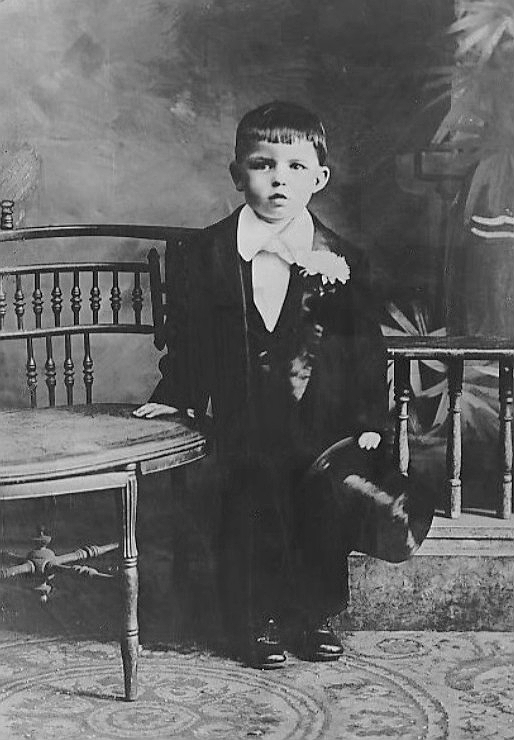 Frank sinatra as a child around the age of 3 in a suit and top hat with his hand on a chair