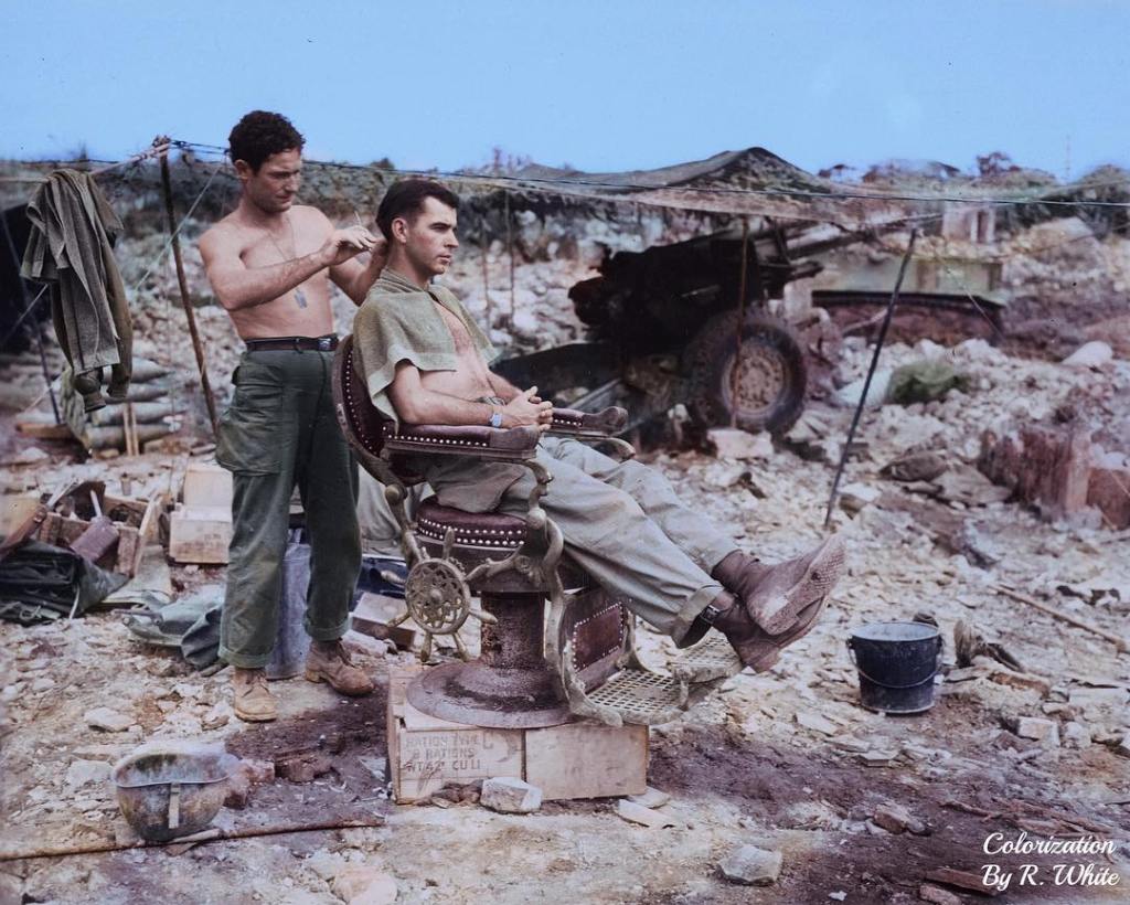 Colorized photograph of Pvt. Troy Dixon (standing) and Sgt. John Anderson (seated), Members of the 363rd Field Artillery Battalion, using a Japanese barber chair to cut hair on 10 June 1945 near Shuri, Okinawa.