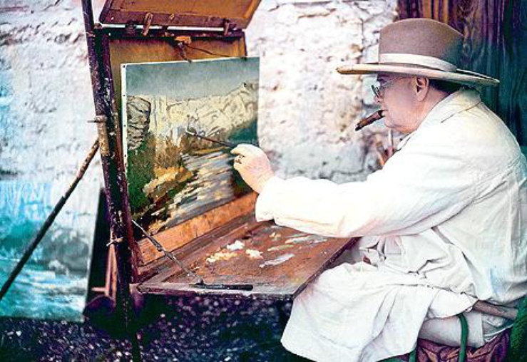 Winston Churchill seated wearing a jacket and large hat, painting a landscape in Southern France in 1955.