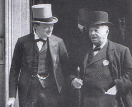 Winston Churchill in a suit and top hat standing on the left of the photo with Lord Jacky Fisher who is also wearing a suit. He has a cane and hat with him. They are standing outside of a building probably in London in 1913.