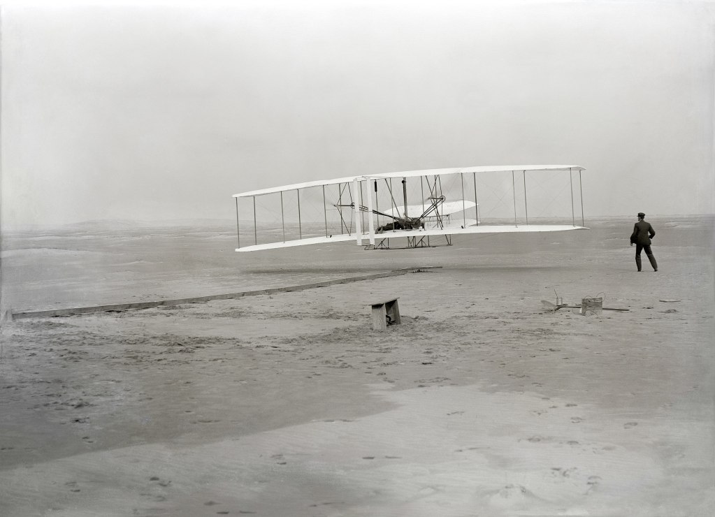 The first powered, controlled, sustained airplane flight in history. Orville Wright, age 32, is at the controls of the machine, lying prone on the lower wing with hips in the cradle which operated the wing-warping mechanism. His brother, Wilbur Wright, age 36, ran alongside to help balance the machine, having just released his hold on the forward upright of the right wing. The starting rail, the wing-rest, a coil box, and other items needed for flight preparation are visible behind the machine.