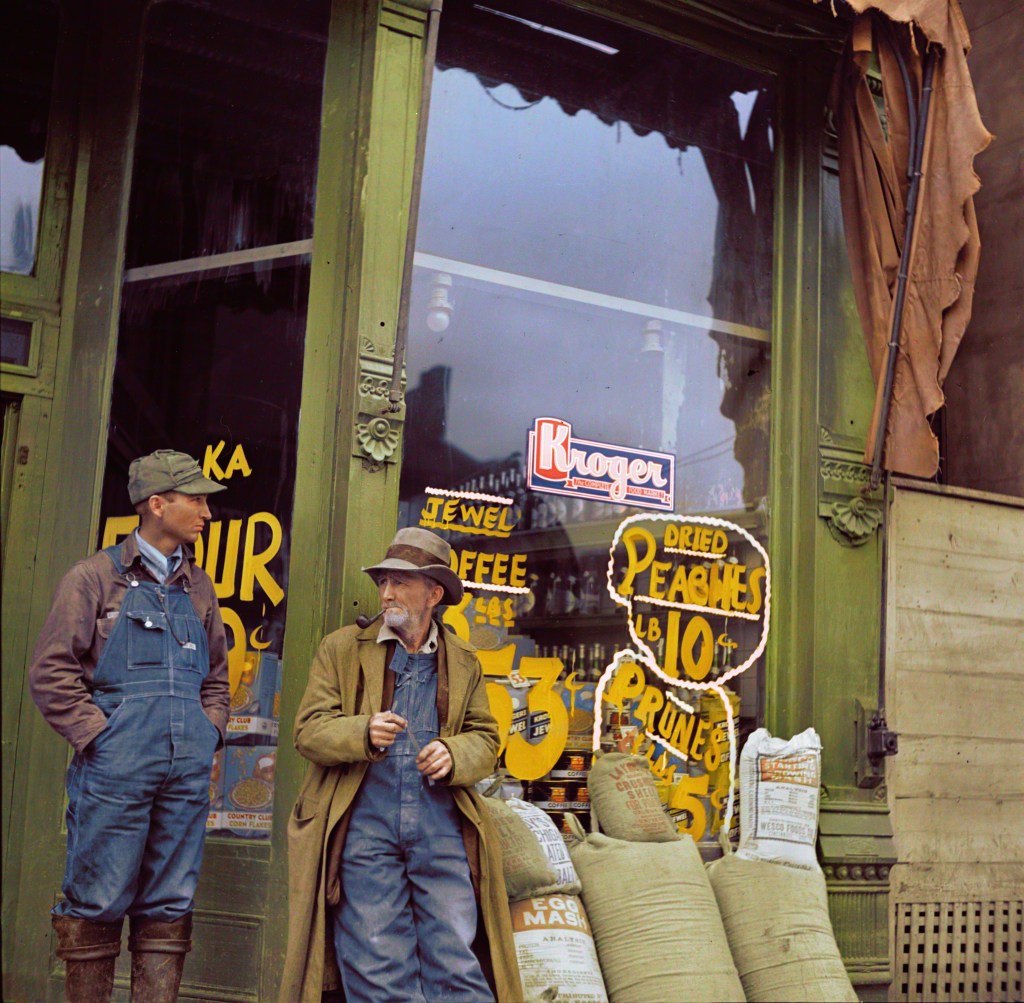 Colorized photograph of Two men standing outside of a grocery store in Shawneetown, Illinois. In the window you can see the price of Dried Peaches, Prunes, and a Kroger sign. In front of the store there are sacks of various products