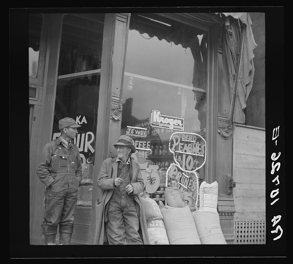 Colorized photograph of Two men standing outside of a grocery store in Shawneetown, Illinois. In the window you can see the price of Dried Peaches, Prunes, and a Kroger sign. In front of the store there are sacks of various products