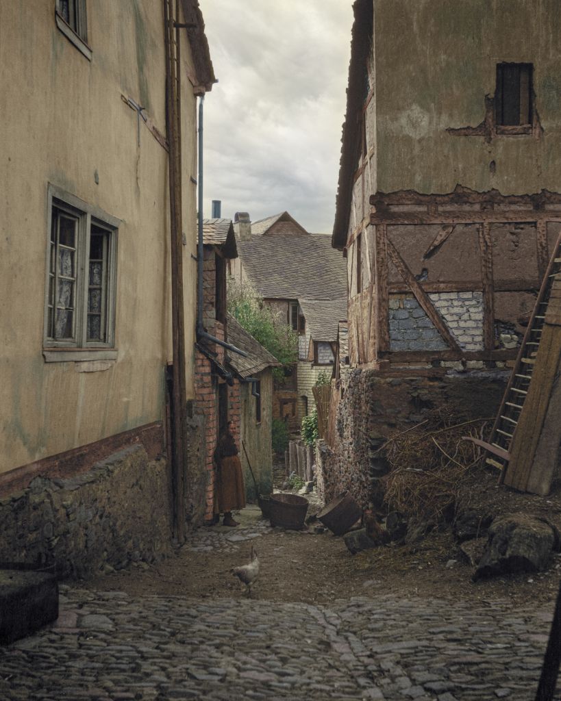 1904 photograph of a woman standing in an alley in an unknown village in Europe, most likely located in Northern France or Belgium. Colorization by Baptiste Pierre