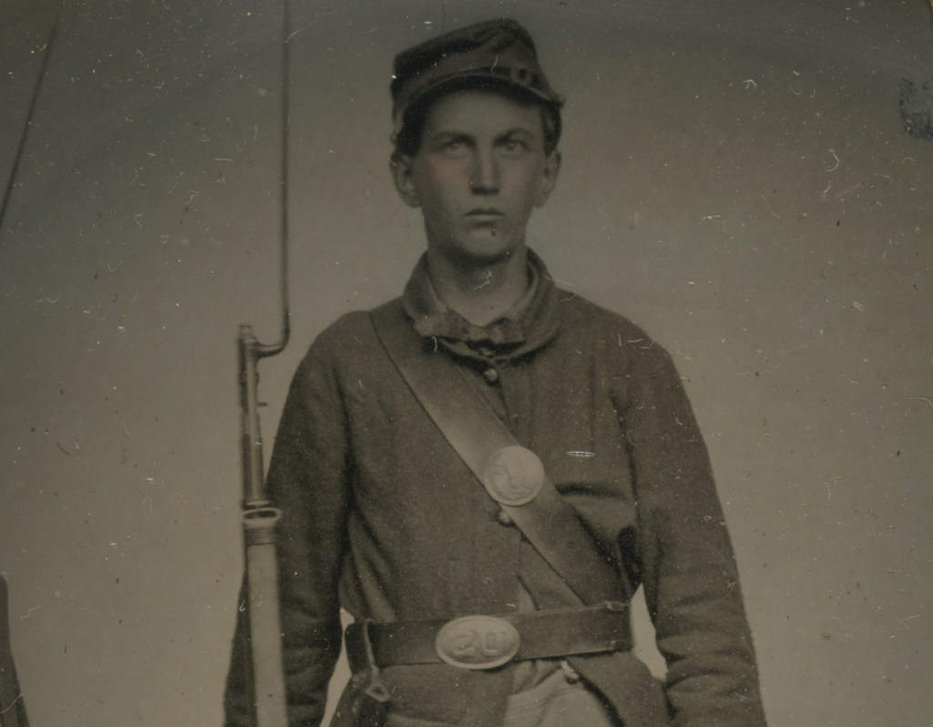 Unidentified young soldier in Union uniform with bayoneted musket next to a rifle stack. photographed during the American Civil War