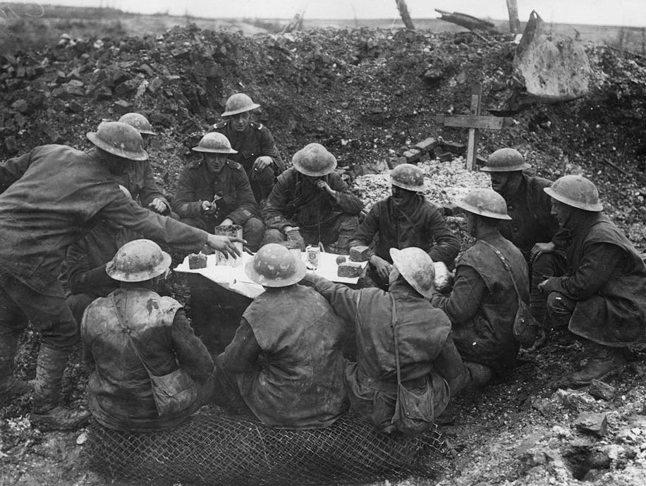 Description from NLS: Soldiers celebrating Christmas in the trenches, Western Front, during World War I. Men sitting in the mud in a trench celebrating Christmas. They have laid some white paper on a crate and set out some slices of cake, a tinned pudding, a packet of raisins and what looks like an orange. These may have been in a food parcel from home, shared by a group of friends.