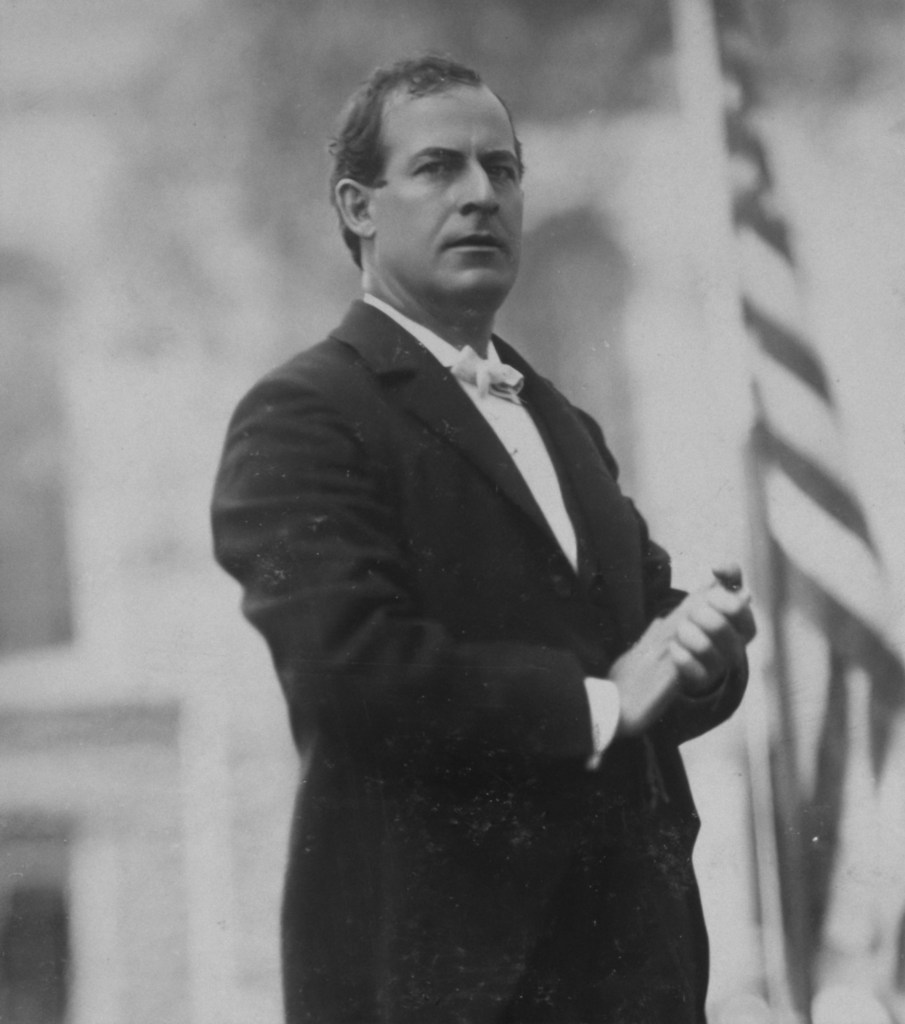 William Jennings Bryan, Democratic party presidential candidate, three-quarter length view standing on stage next to American flag. Photograph taken in 1896 during the Presidential election campaign