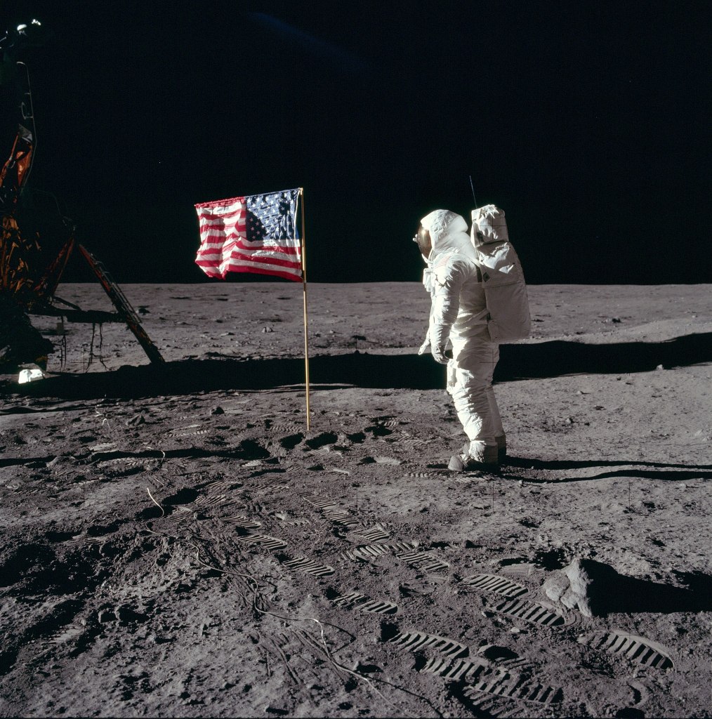 "Buzz Aldrin salutes the U.S flag on the Moon (mission time: 110:10:33). His fingertips are visible on the far side of his faceplate. Note the well-defined footprints in the foreground. Buzz is facing up-Sun. There is a reflection of the Sun in his visor. At the bottom of Buzz's faceplate, note the white 'rim' which is slightly separated from his neckring. This 'rim' is the bottom of his gold visor, which he has pulled down. We can see the LEC straps hanging down inside of the ladder strut. In the foreground, we can see the foot-grabbing loops in the TV cable. The double crater under Neil's LM window is just beyond the LM shadow." via Wikimedia Commons